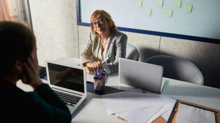 Managing director sitting in front of whiteboard with green marker and looking at another person at her desk