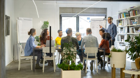 Confident mature businessman giving a presentation to his crative team in office. Business brief with annual goals with casual employees. Happy leadership man training young businessmen and smiling businesswomen sitting at conference table.